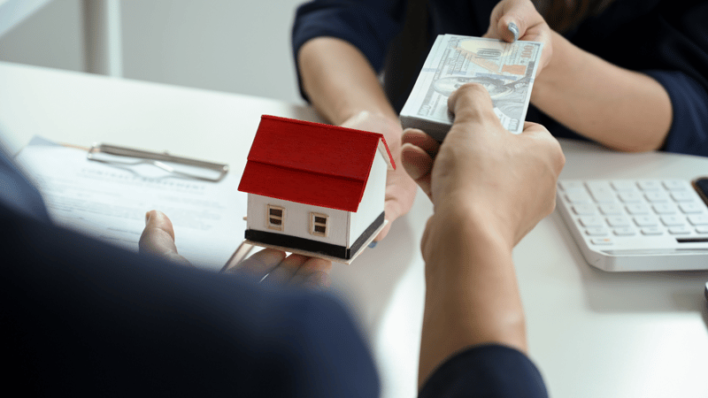 Two people exchanging cash over a desk with a small red-roofed model house