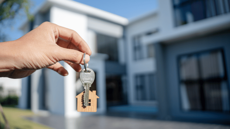 Hand holding house keys with a house-shaped keychain in front of a modern home