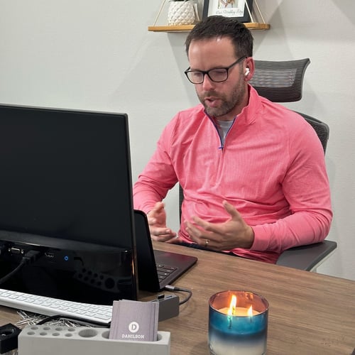Man in a pink shirt talking at a desk in front of a computer monitor
