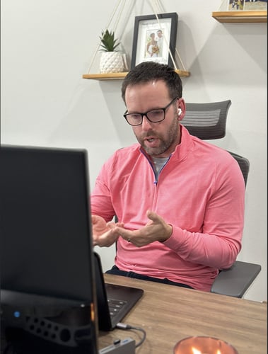 Man in a pink shirt talking at a desk in front of a computer monitor