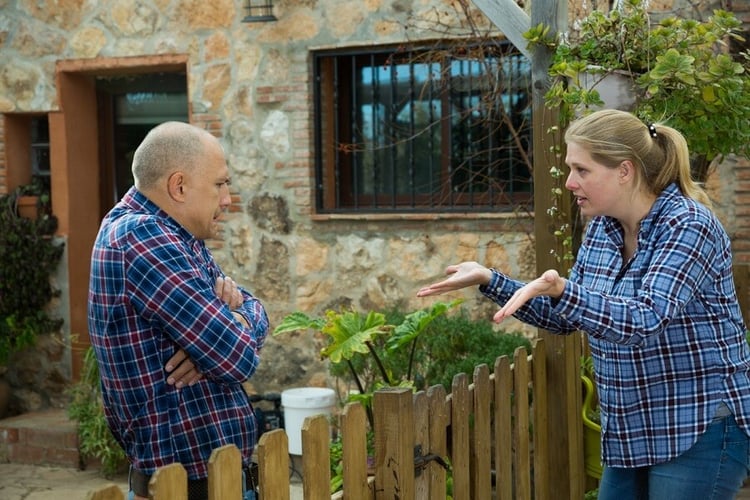 Two neighbors talking across a wooden fence outside a stone house