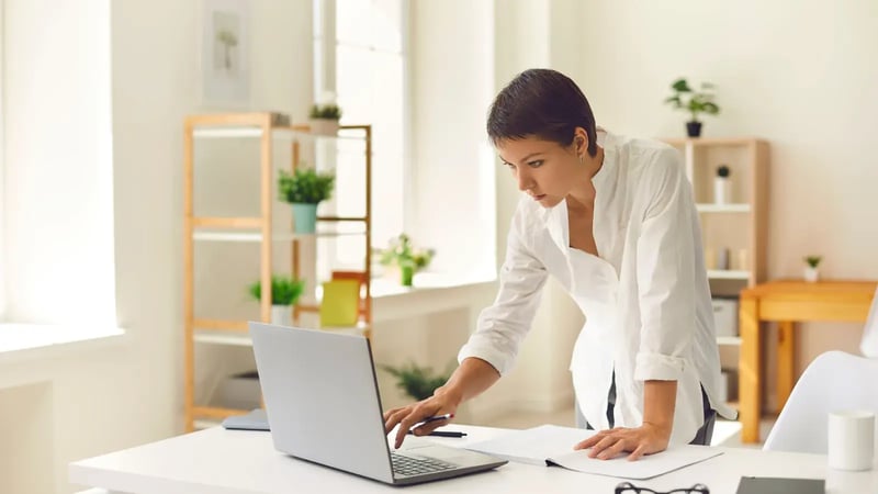Woman standing at a desk typing on a laptop in a bright home office