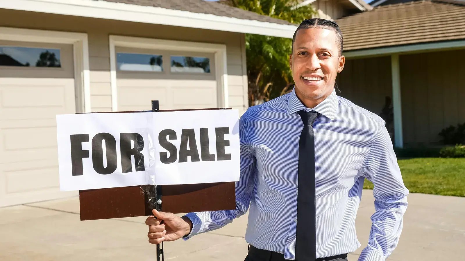 Agent in a shirt and tie holding a For Sale sign in front of a house