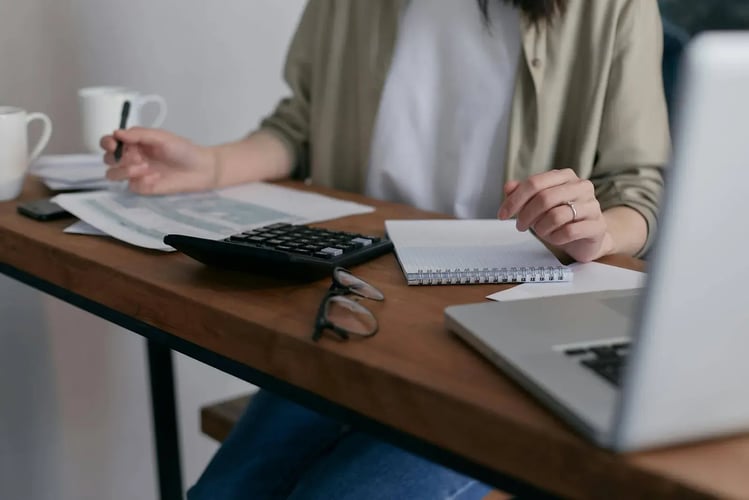 Person at a desk with a laptop, calculator and notebook, holding a mug