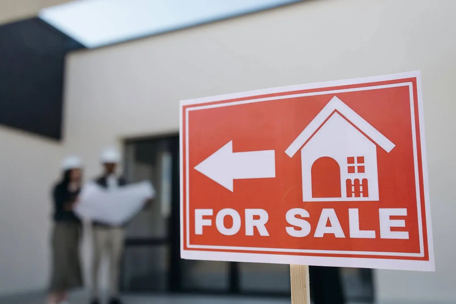 Red For Sale sign with a house icon in front of a home, people reviewing papers behind it