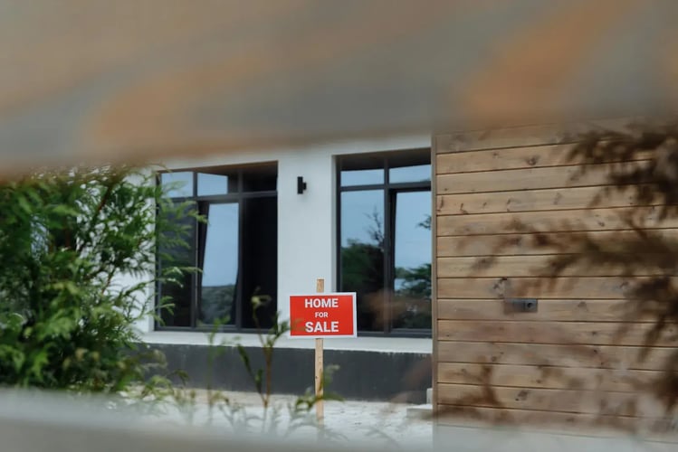 Modern house exterior with a red Home For Sale sign out front, seen through foliage