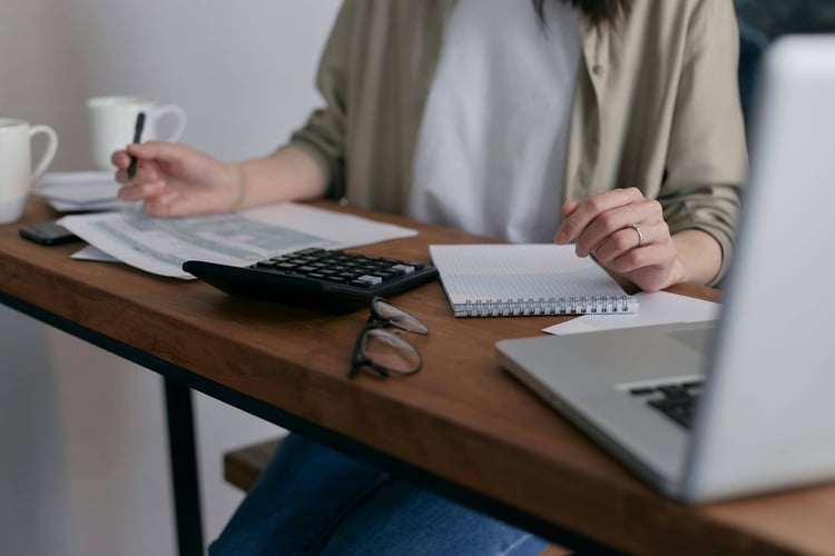 Person at a desk with a laptop, calculator and notebook, holding a mug